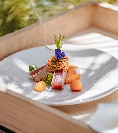 Gourmeturlaub Ein kunstvoll angerichteter Teller mit frischem Fisch, buntem Gemüse und einer essbaren Blüte im Design Hotel Sonnen Resort Naturns.